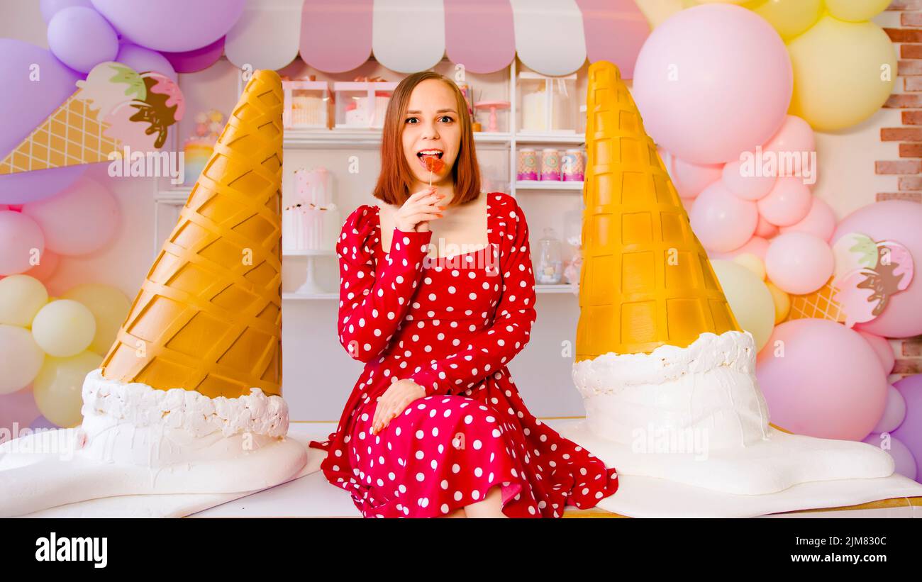 Contenido femenino mirando la cámara mientras come chuleta dulce sobre la mesa con grandes conos decorativos de helado en confitería ligera Foto de stock