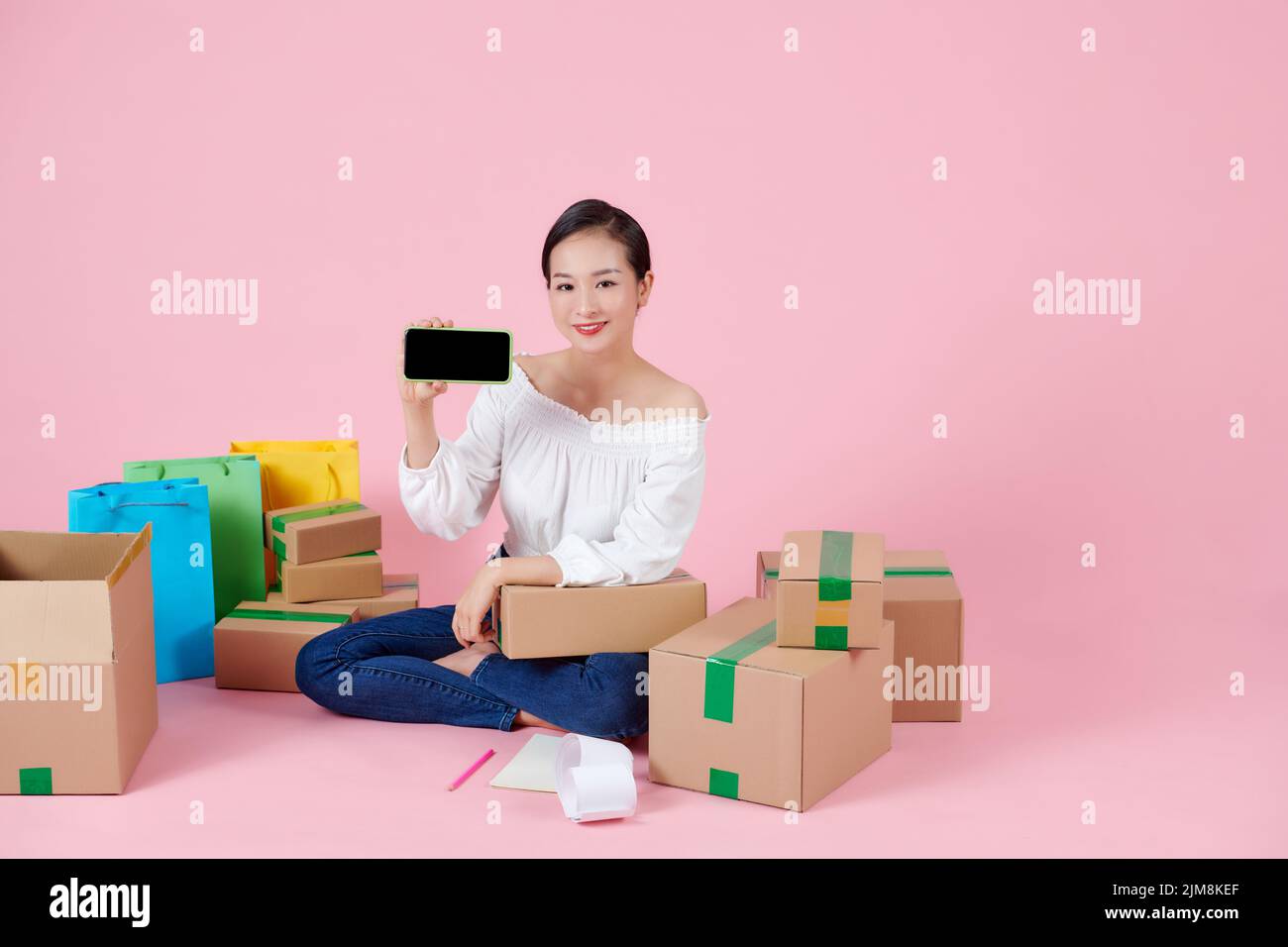 Mujer feliz mostrando smartphone con pantalla en blanco negro, sentado entre cajas después de moverse, la publicidad de la agencia inmobiliaria Foto de stock