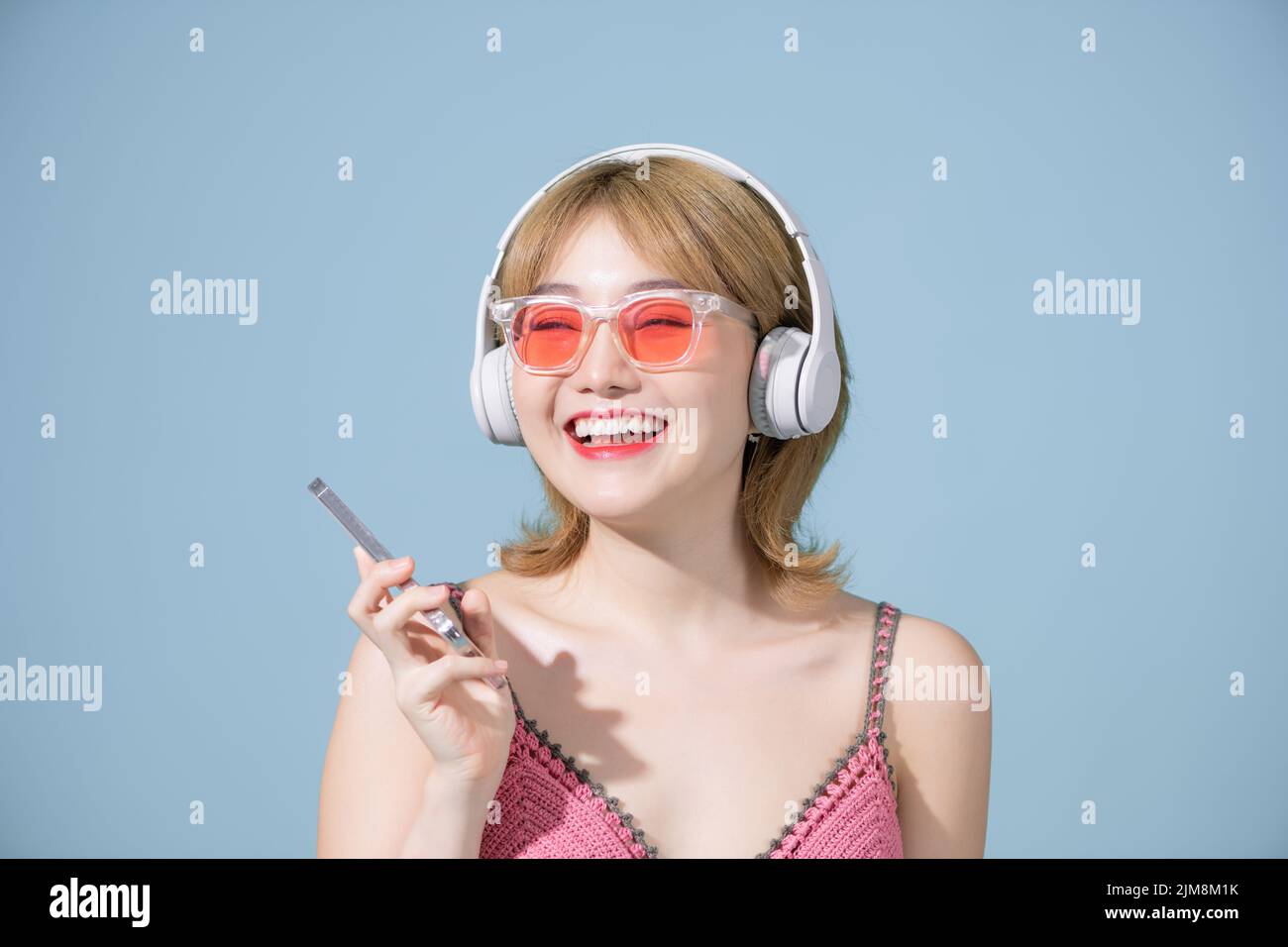 La fotografía de estudio de una feliz chica asiática del milenio sonríe con alegría utiliza el smartphone y los auriculares Foto de stock