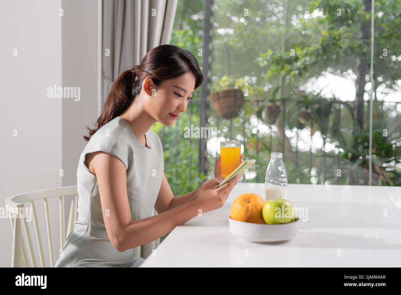 Mujer sonriente y bonita mirando al teléfono móvil y sosteniendo un vaso de zumo de naranja mientras desayunaba Foto de stock