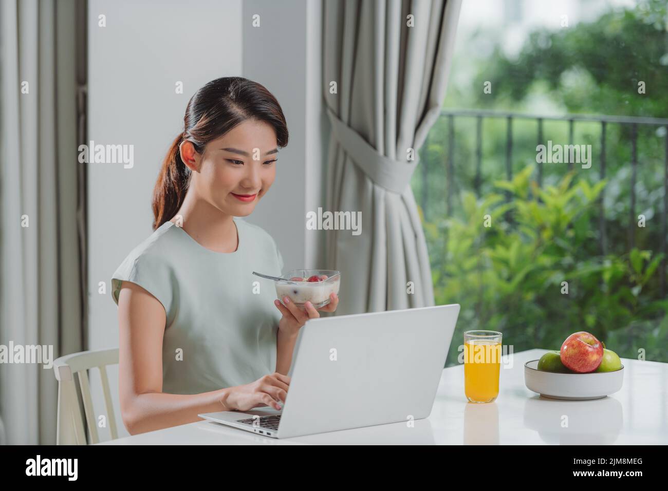 Retrato de una hermosa joven que trabaja con el ordenador portátil mientras come en casa. Foto de stock