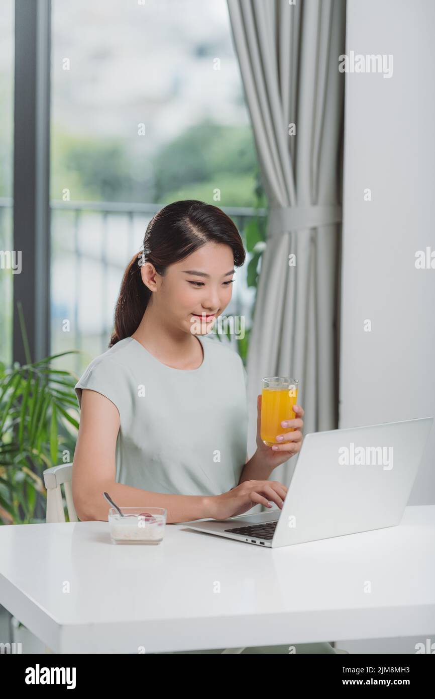 hermosa joven que trabaja con el ordenador portátil mientras desayuna con cereales y leche y bebe zumo de naranja Foto de stock