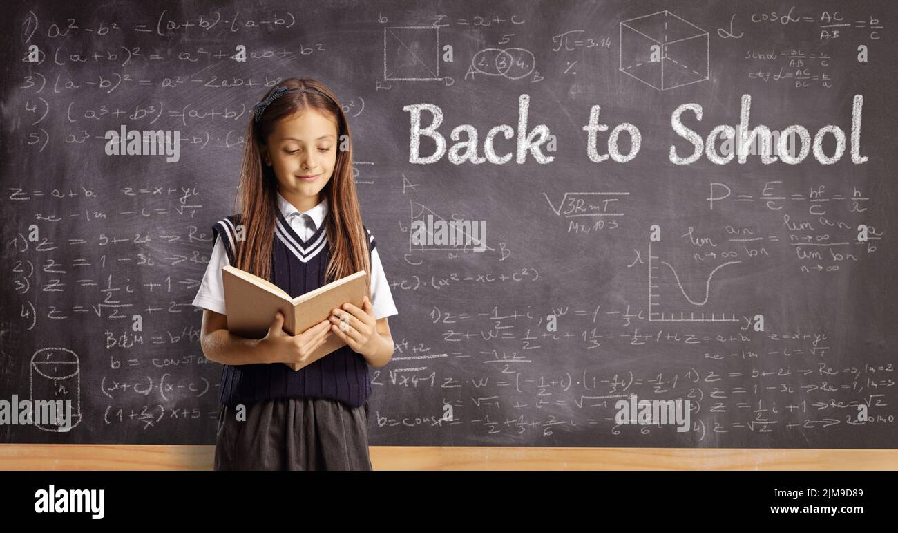 Alumna leyendo un libro delante de una pizarra con fórmulas de matemáticas y texto de vuelta a la escuela Foto de stock