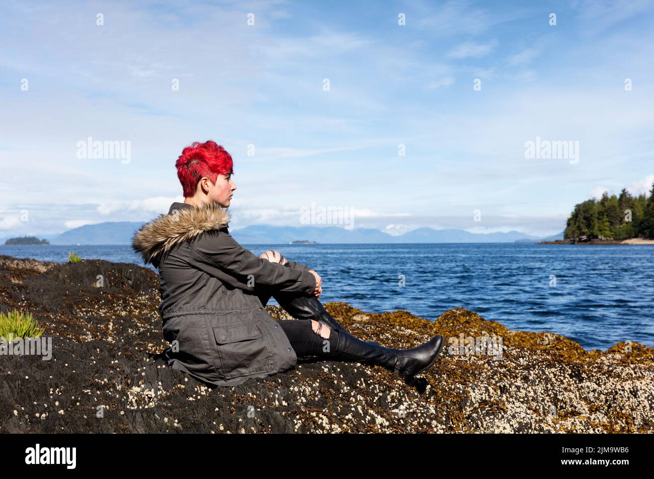 Niña adolescente pensando mientras se sienta en la roca cerca de lago y bosques Foto de stock