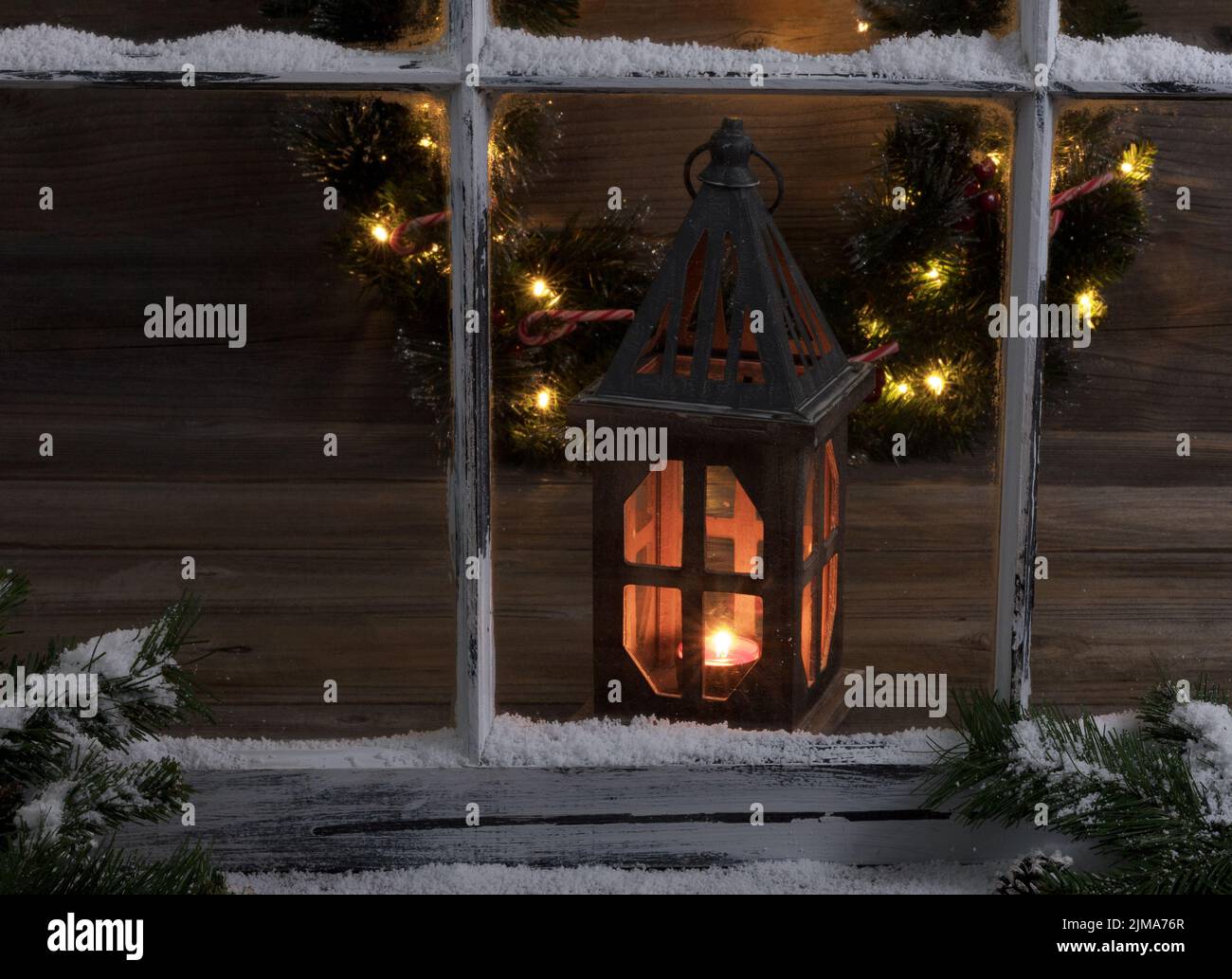 Ventana nevada y ramas de abeto con linterna brillante y la corona de Navidad de luz Foto de stock