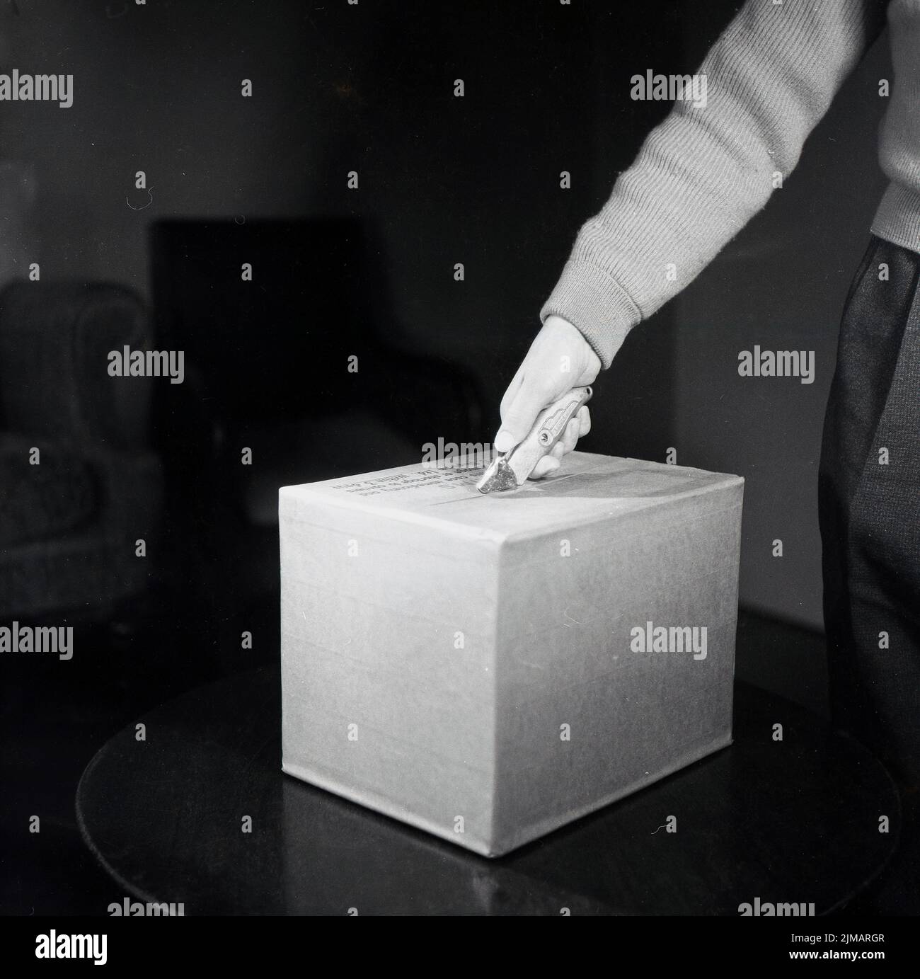 1950s, histórico, un hombre - con la mano y el antebrazo en el tiro - demostrando cómo abrir una caja de cartón con una herramienta de hoja de mango metálico, fabricada por Lewis. Este cuchillo de uso general era conocido como un 'cortador de cajas'. Foto de stock