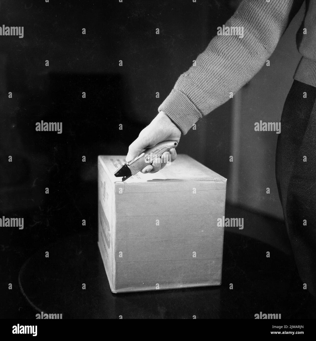 1950s, histórico, un hombre - con la mano y el antebrazo en el tiro - demostrando cómo abrir una caja de cartón con una herramienta de hoja de mango metálico, fabricada por Lewis. Este cuchillo de uso general era conocido como un 'cortador de cajas'. Foto de stock