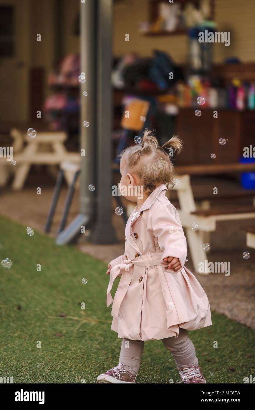 Niña pequeña con pigtails en el centro de cuidado diurno usando lindo abrigo rosa rodeado de burbujas, sin rostro Foto de stock