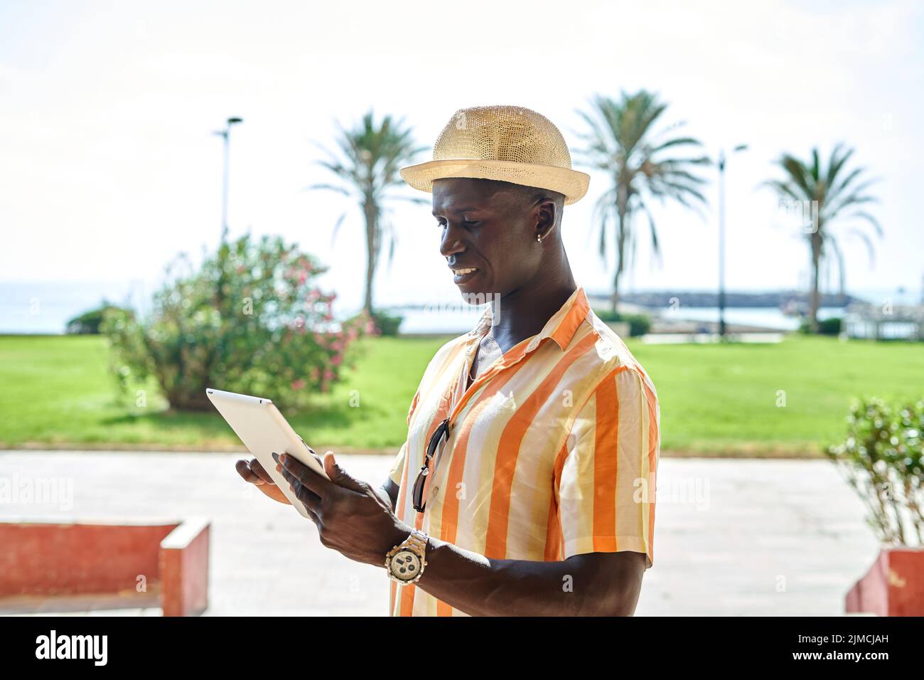 Vista lateral de la tableta de surf de hombre de negocios afroamericano mientras se encuentra en la costa y el mar en el soleado día de verano en un complejo tropical Foto de stock