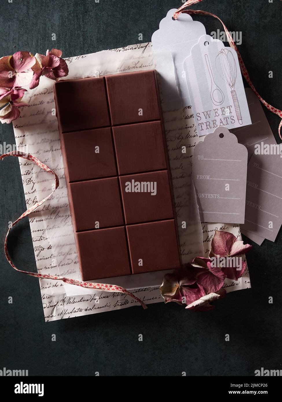 Vista superior de la sabrosa barra de chocolate dulce en envoltorio de regalo con flores secas y etiquetas sobre fondo oscuro en la habitación Foto de stock