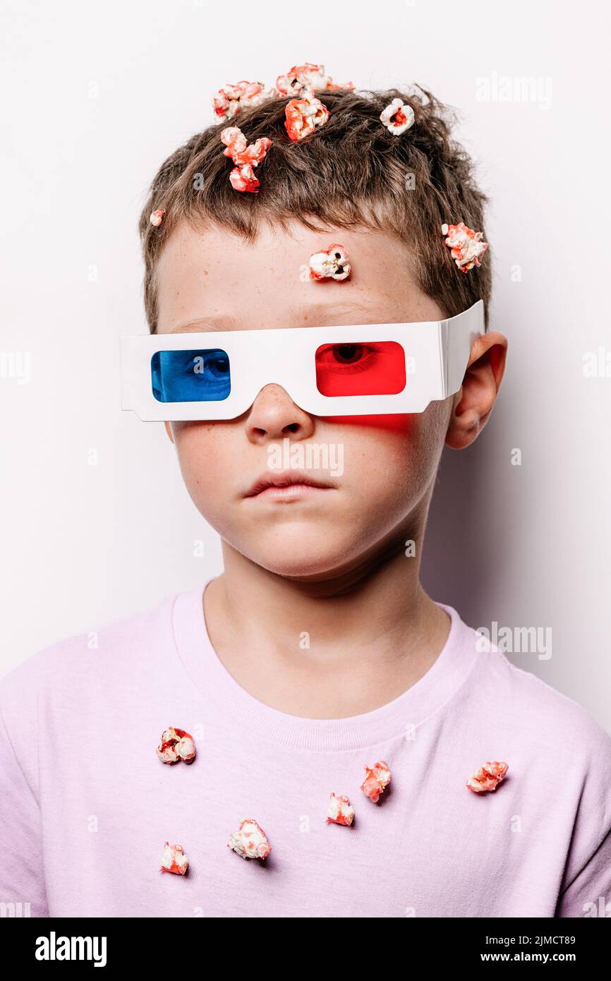 Lindo niño en gafas 3D con lentes de colores lanzando palomitas de maíz sobre la cabeza mientras mira la cámara sobre fondo blanco en un estudio de luz Foto de stock