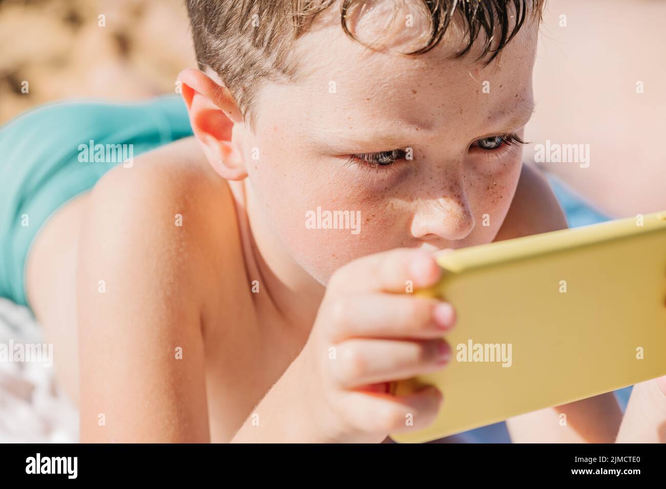 Niño centrado en nadar pantalones cortos jugando a videojuegos en el teléfono móvil mientras yacía en la orilla arenosa en un complejo tropical en un soleado día de verano en Getaria Zarautz Foto de stock