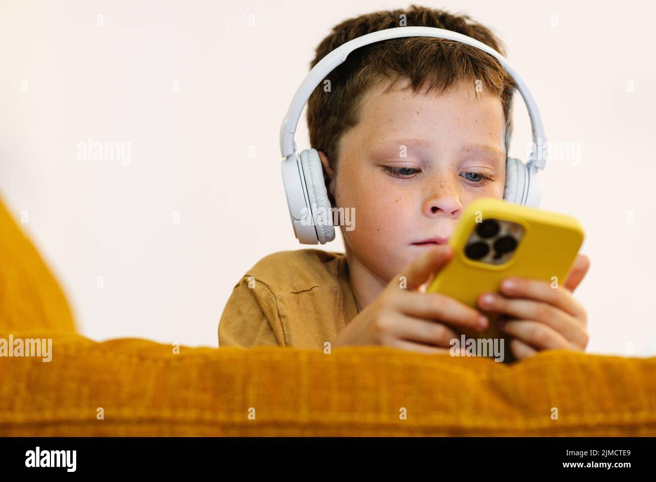 Niño centrado tumbado en un sofá escuchando música con los auriculares en la cabeza y utilizando el teléfono móvil Foto de stock