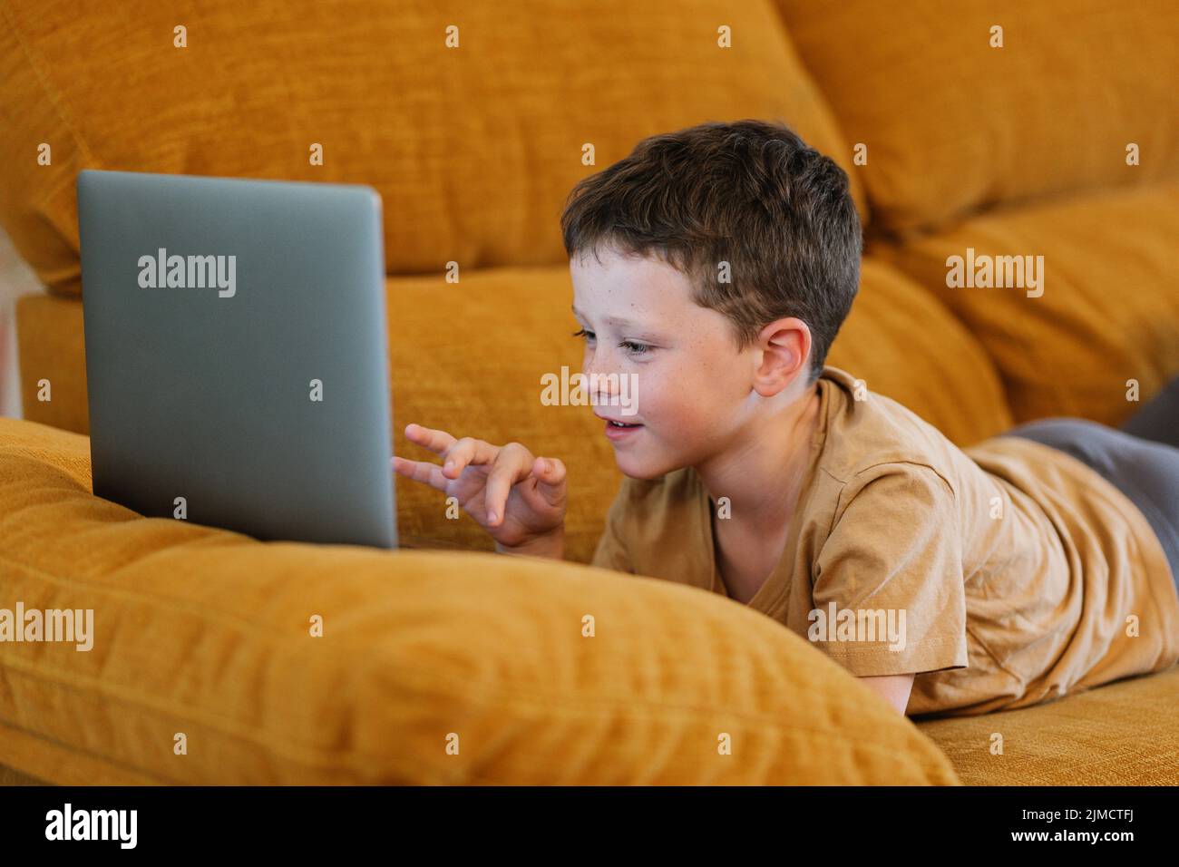Vista lateral del niño feliz tumbado en un sofá con el ordenador portátil mientras está en casa Foto de stock