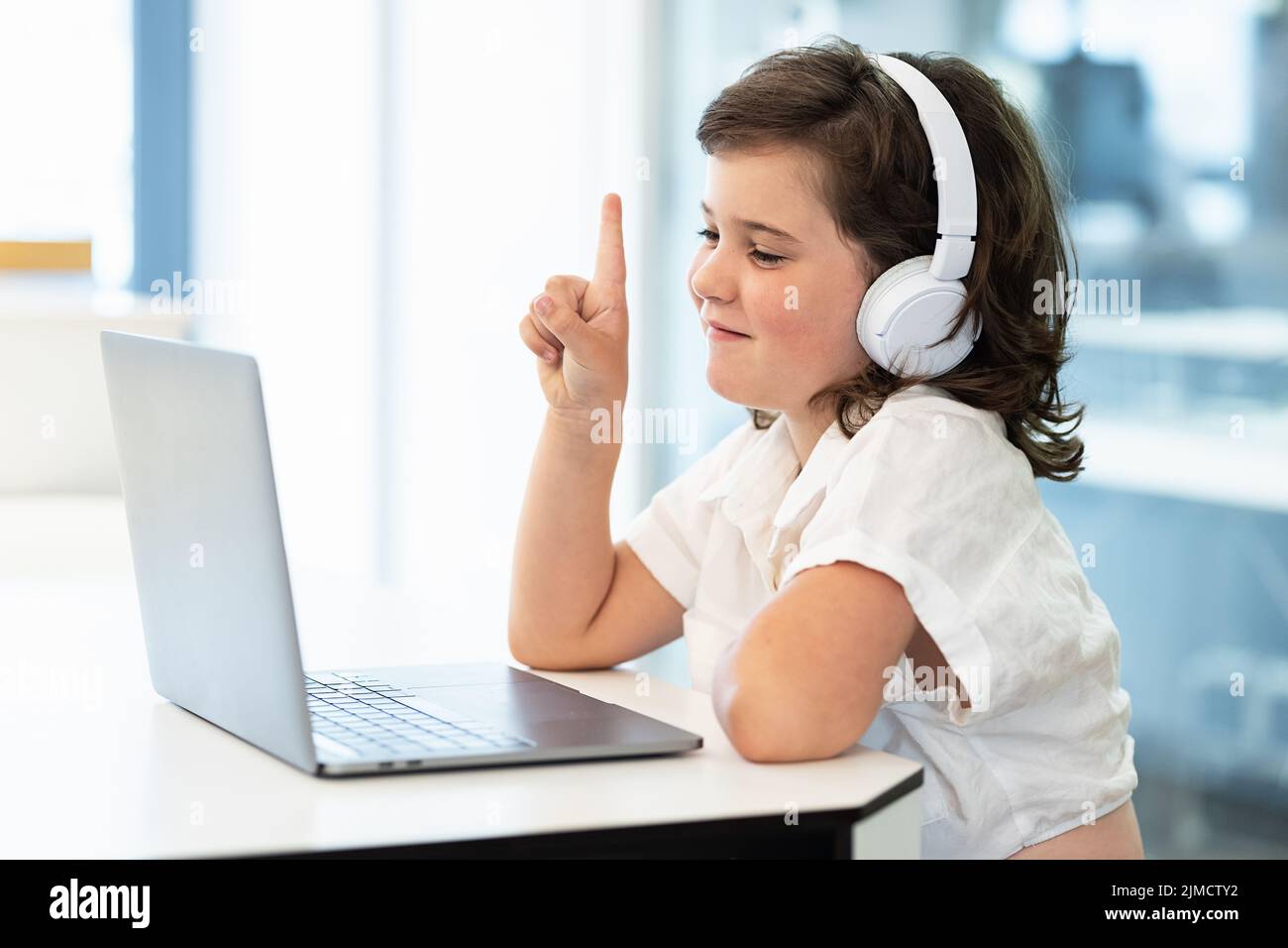 Vista lateral de la chica con auriculares en la cabeza sentada frente a un portátil durante el chat de vídeo despertando en la pantalla Foto de stock