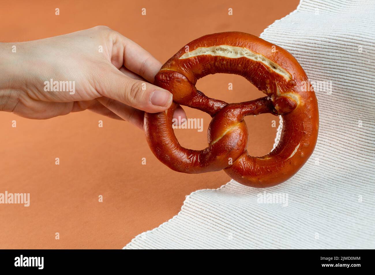 Panadero femenino irreconocible que muestra un delicioso pretzel recién horneado con corteza oscura servido sobre tejido estriado sobre fondo marrón Foto de stock
