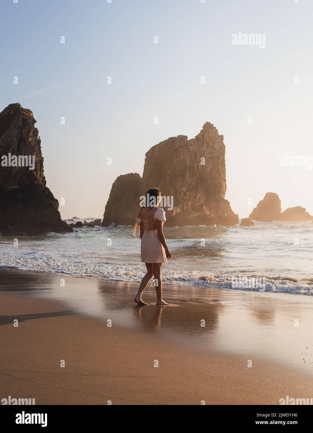Atrás ver mujer viajero caminando en la costa arenosa con acantilados rocosos ásperos cerca del mar en la puesta de sol en la soleada noche de verano Foto de stock
