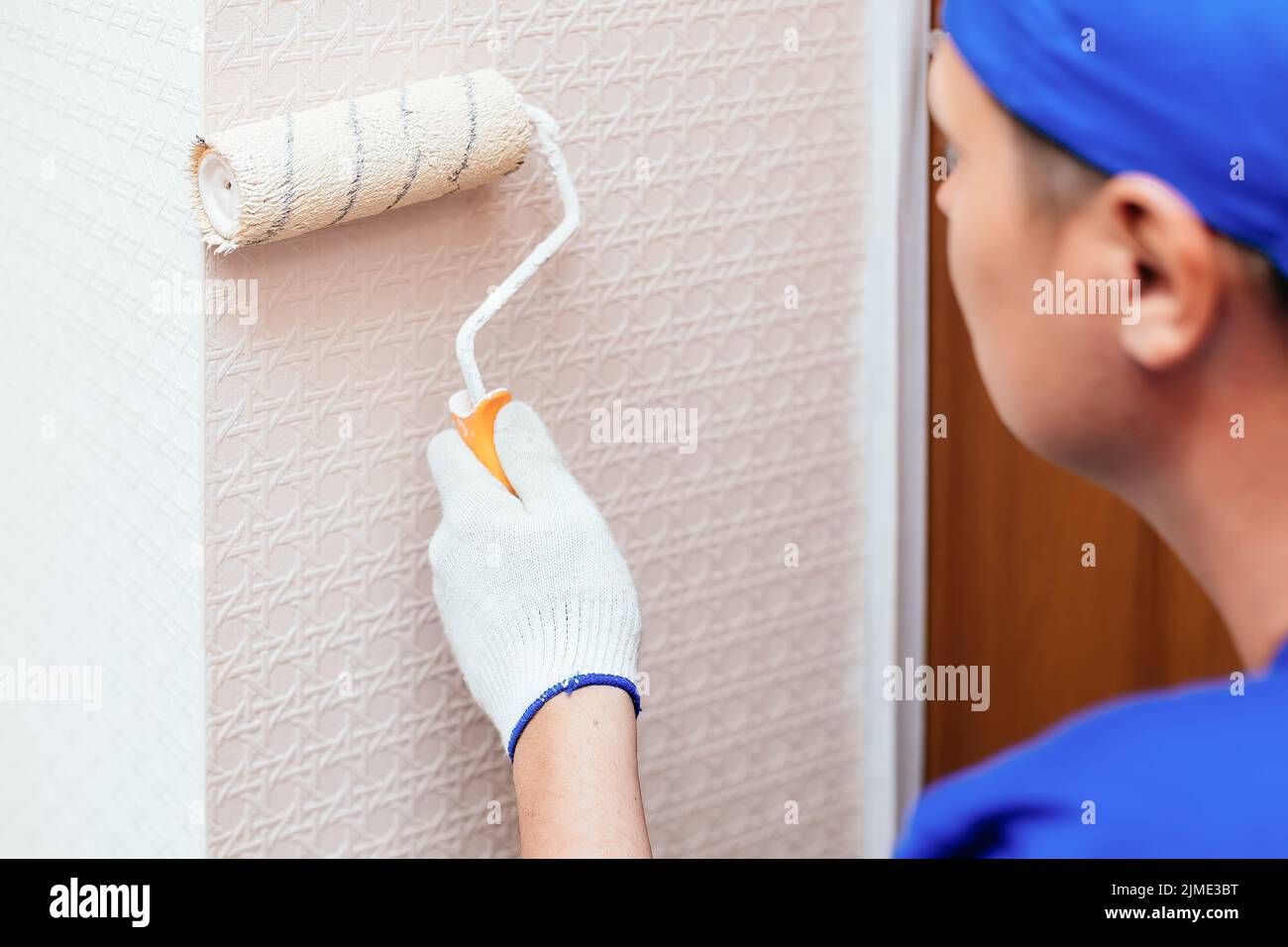 Un trabajador de apariencia caucásica hace reparaciones al apartamento y pinta la pared con un rodillo de pintura en color durazno. Foto de stock