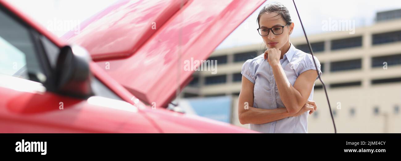 Una mujer joven mira la capucha abierta de un coche rojo Foto de stock