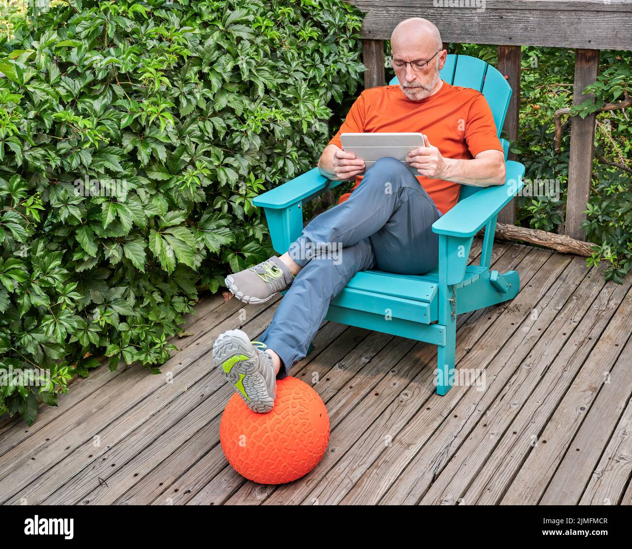 El anciano con una tableta digital se está relajando en una silla Adirondack en una terraza de madera en el patio trasero Foto de stock