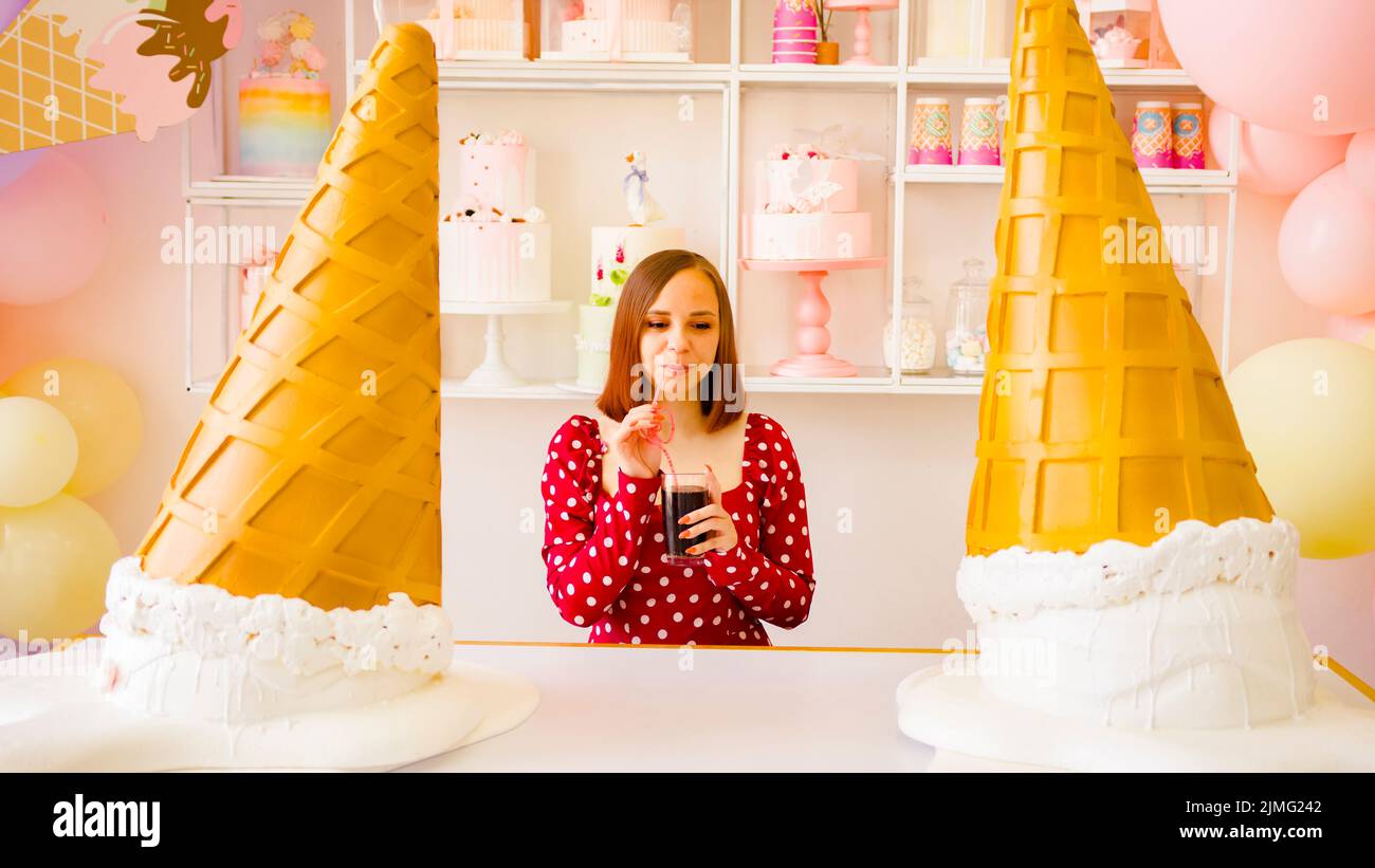 Mujer de ensueño bebiendo bebida refrescante cerca de grandes conos decorativos de helado. Foto de stock
