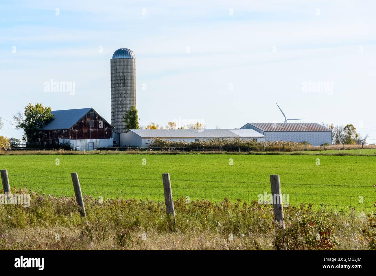 Antiguo granero de metal y silo en una granja en el extremo de un campo de hierba en un soleado día de otoño. Un aerogenerador es visible en el fondo. Foto de stock