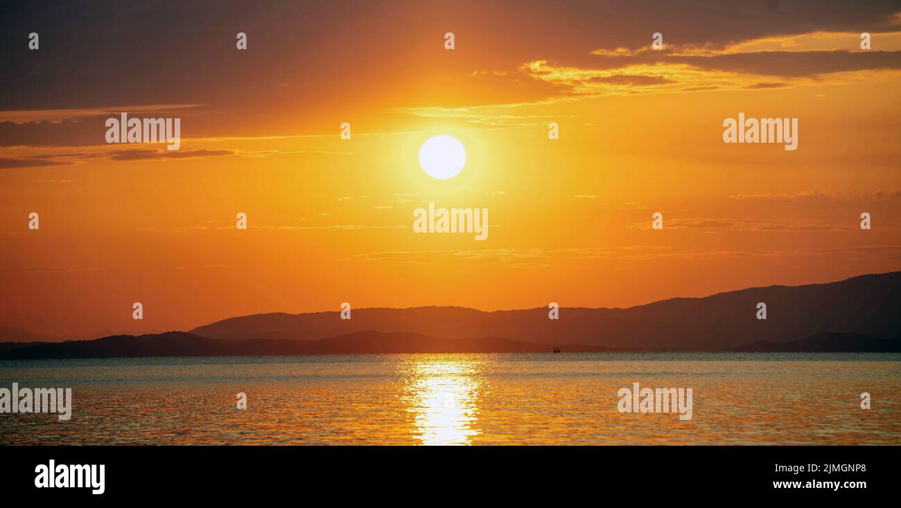 Puesta de sol sobre el mar Egeo. Grecia. Sol cayendo detrás de tierra oscura, reflejos dorados sobre el agua ondulada del océano. Color naranja cielo y nubes Foto de stock