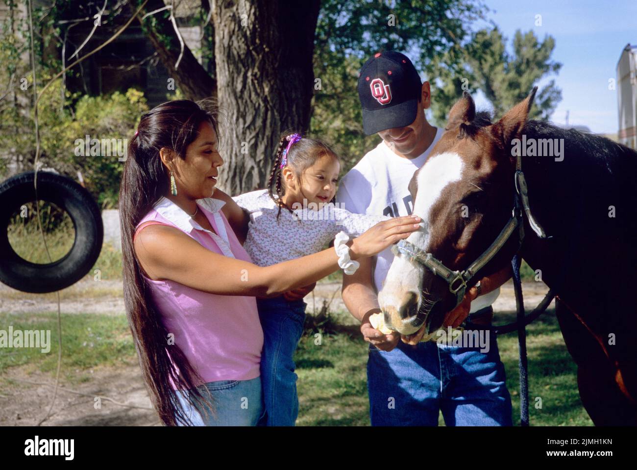 La familia nativa americana de madre y padre muestra a su hija de 4 años cómo acariciar con seguridad la cabeza de un caballo Foto de stock