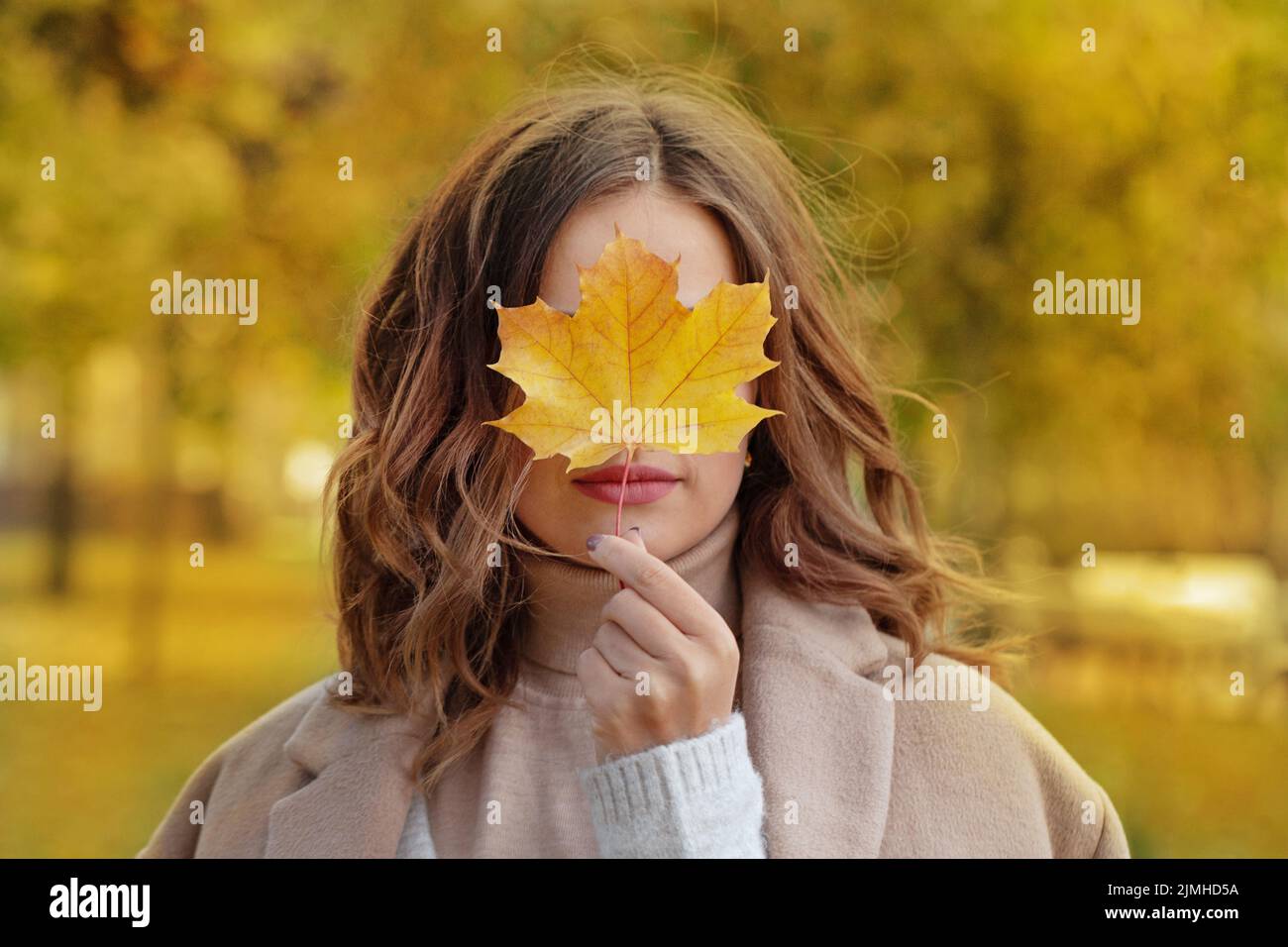 Hermosa mujer sonriente sosteniendo la hoja de arce amarillo otoño cubriendo sus ojos en el parque de otoño Foto de stock