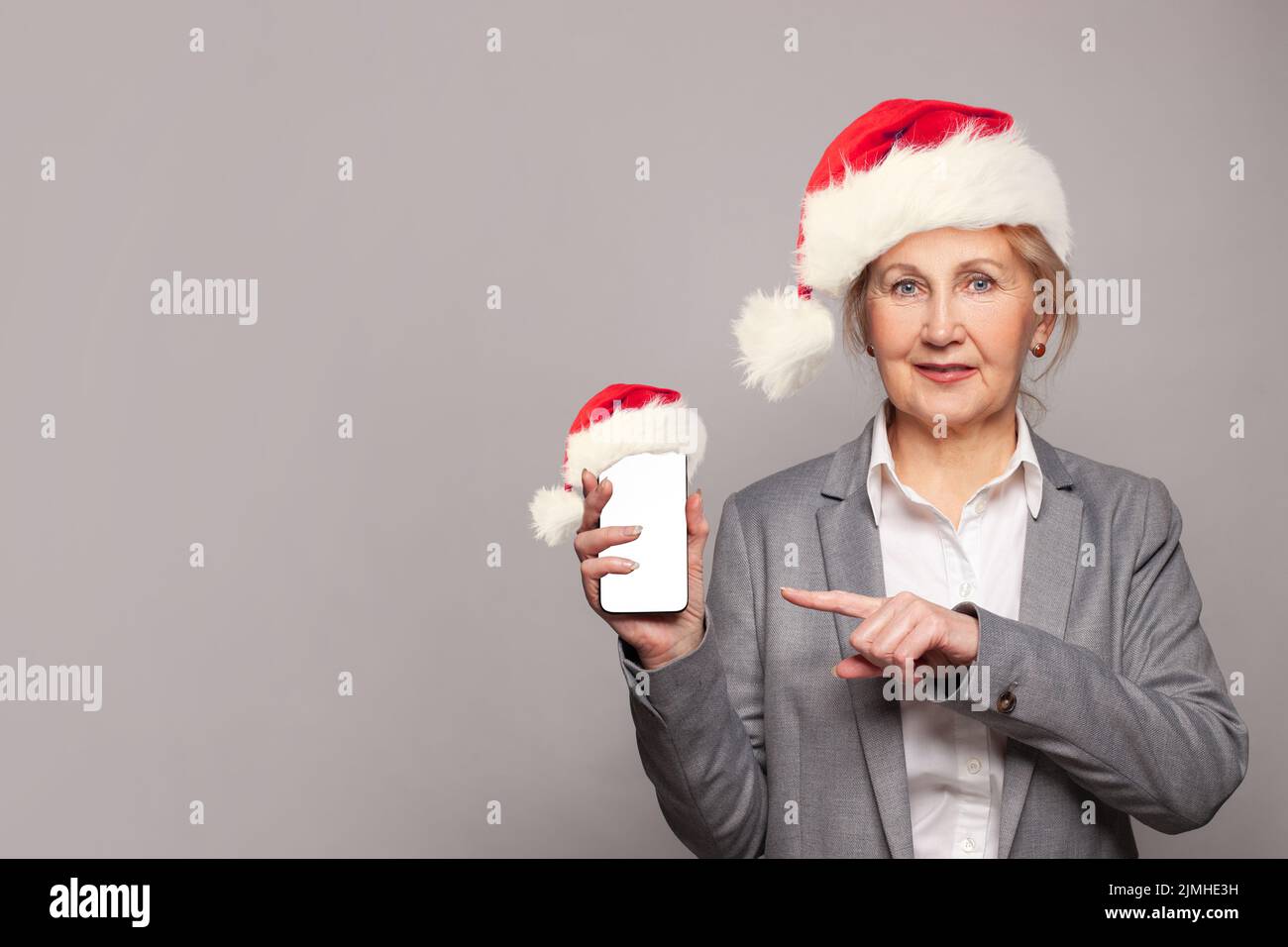 Mujer mayor de Navidad con sombrero de Papá Noel con smartphone Papá Noel con pantalla vacía en blanco sobre el fondo gris de la pancarta de la pared Foto de stock