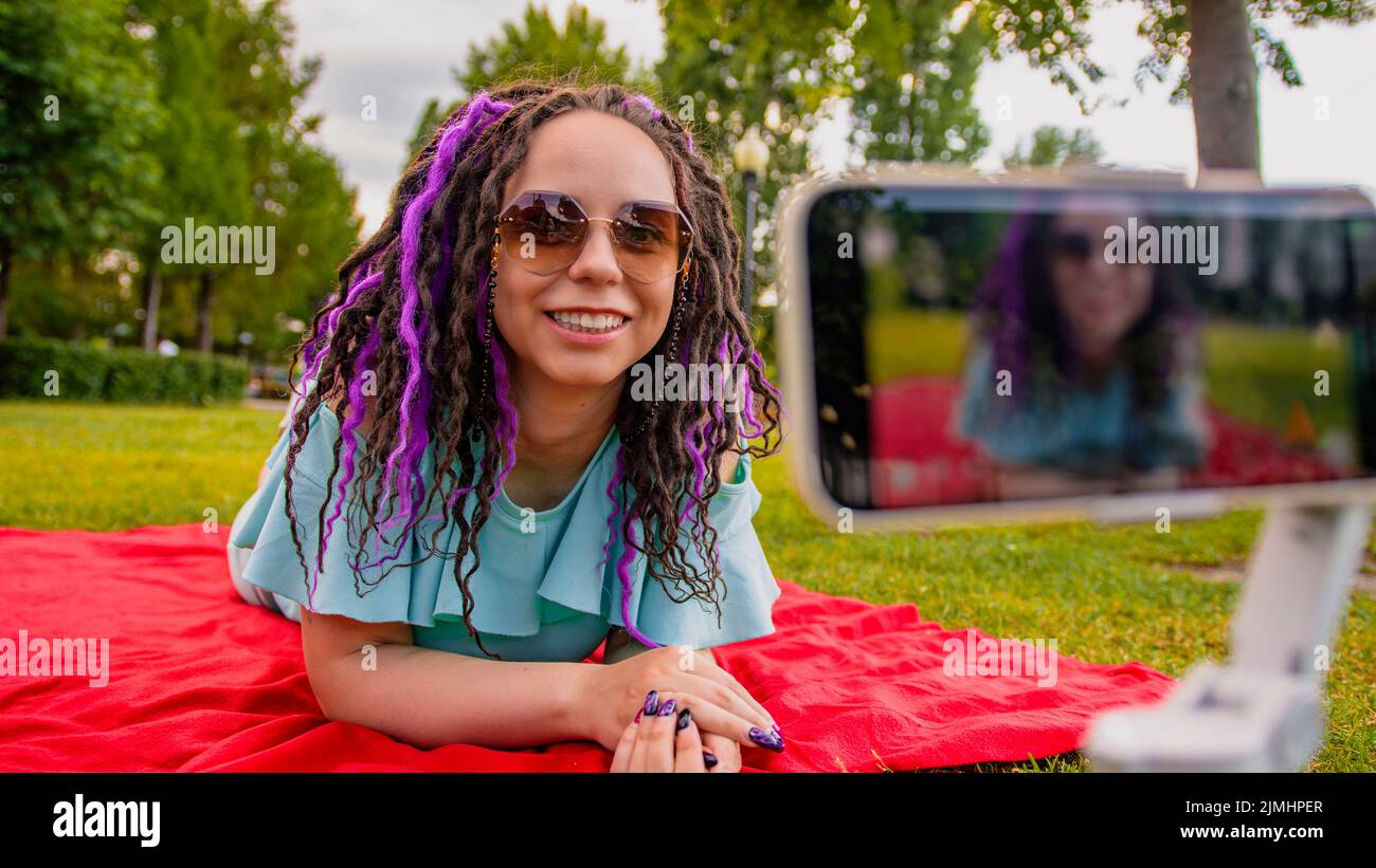 Una mujer con un estilo de peinado descansa sobre una manta en el parque y graba un blog de vídeo con su smartphone. Un blogger hembra dispara un vídeo en una cámara Foto de stock