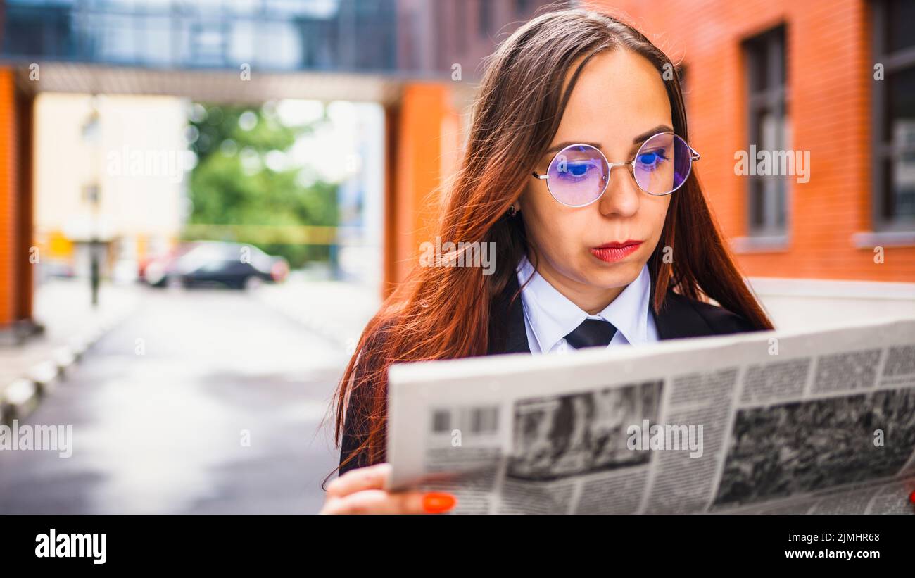 Joven pensativa leyendo el periódico en la calle. Mujer joven empresaria confundida con pelo largo marrón en ropa formal y gafas de pie Foto de stock