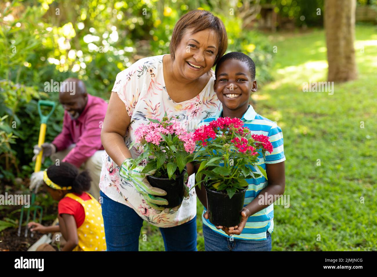 Retrato de niño y abuela afroamericano feliz llevando plantas florecientes en macetas en el patio trasero. Inalterado, familia, unión, fin de semana, pasatiempos Foto de stock