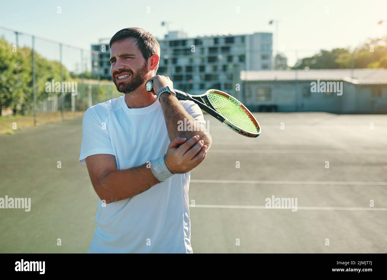 El codo de tenista puede ser causado por movimientos repetitivos de muñeca y brazo. Un hombre joven y deportivo que sostiene el codo con dolor mientras juega al tenis en una cancha de tenis. Foto de stock