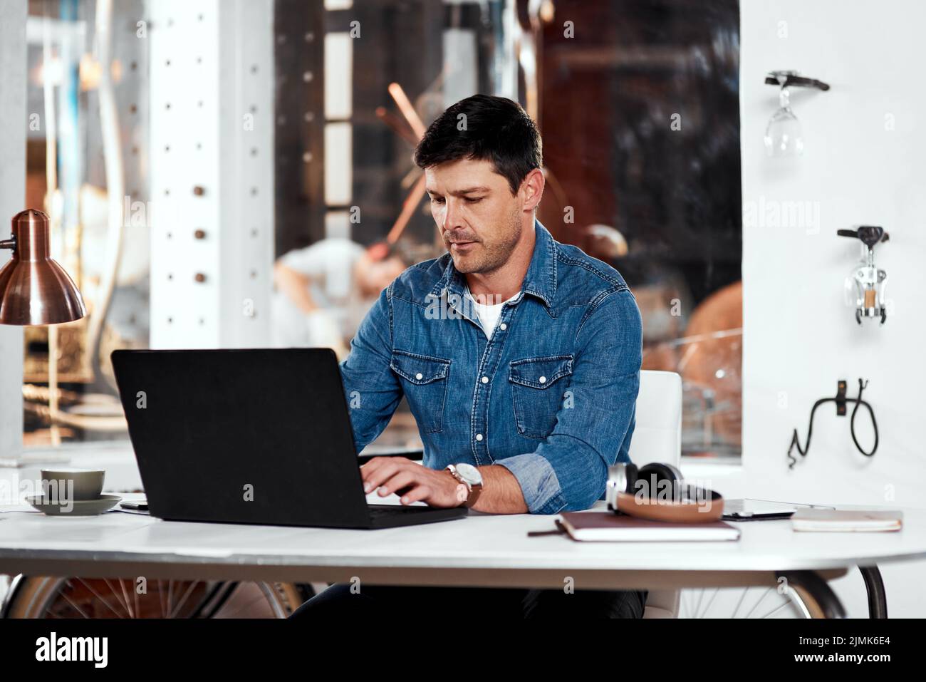 Trabajar duro es una segunda naturaleza para él. Un joven hombre de negocios guapo que trabaja con un ordenador portátil en una oficina dentro de su taller. Foto de stock