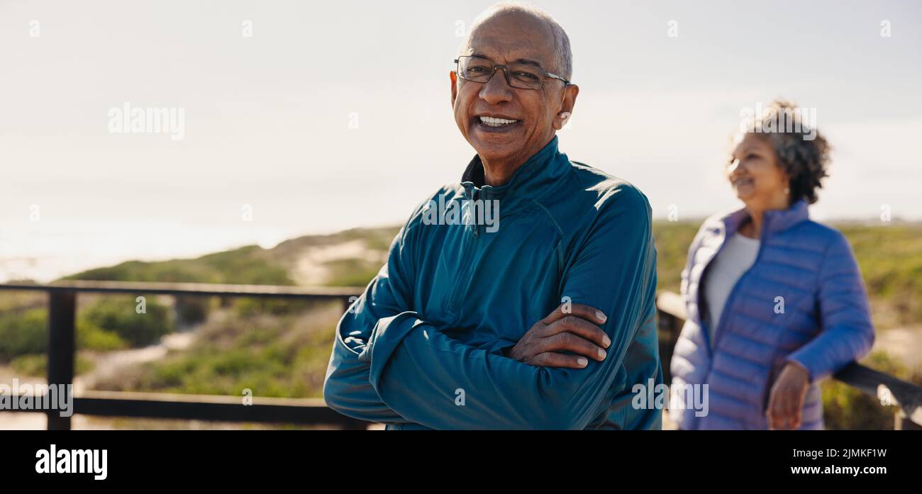 Hombre anciano alegre sonriendo a la cámara mientras estaba de pie en un paseo marítimo con los brazos cruzados. Hombre mayor feliz disfrutando de unas vacaciones relajantes junto al mar w Foto de stock