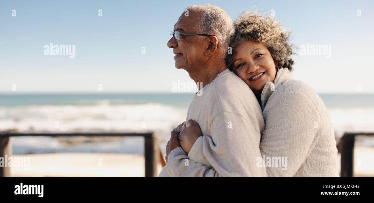 Romántica mujer mayor sonriendo a la cámara mientras abrazaba a su marido junto al océano. Pareja de ancianos afectuosa disfrutando pasar tiempo de calidad Foto de stock