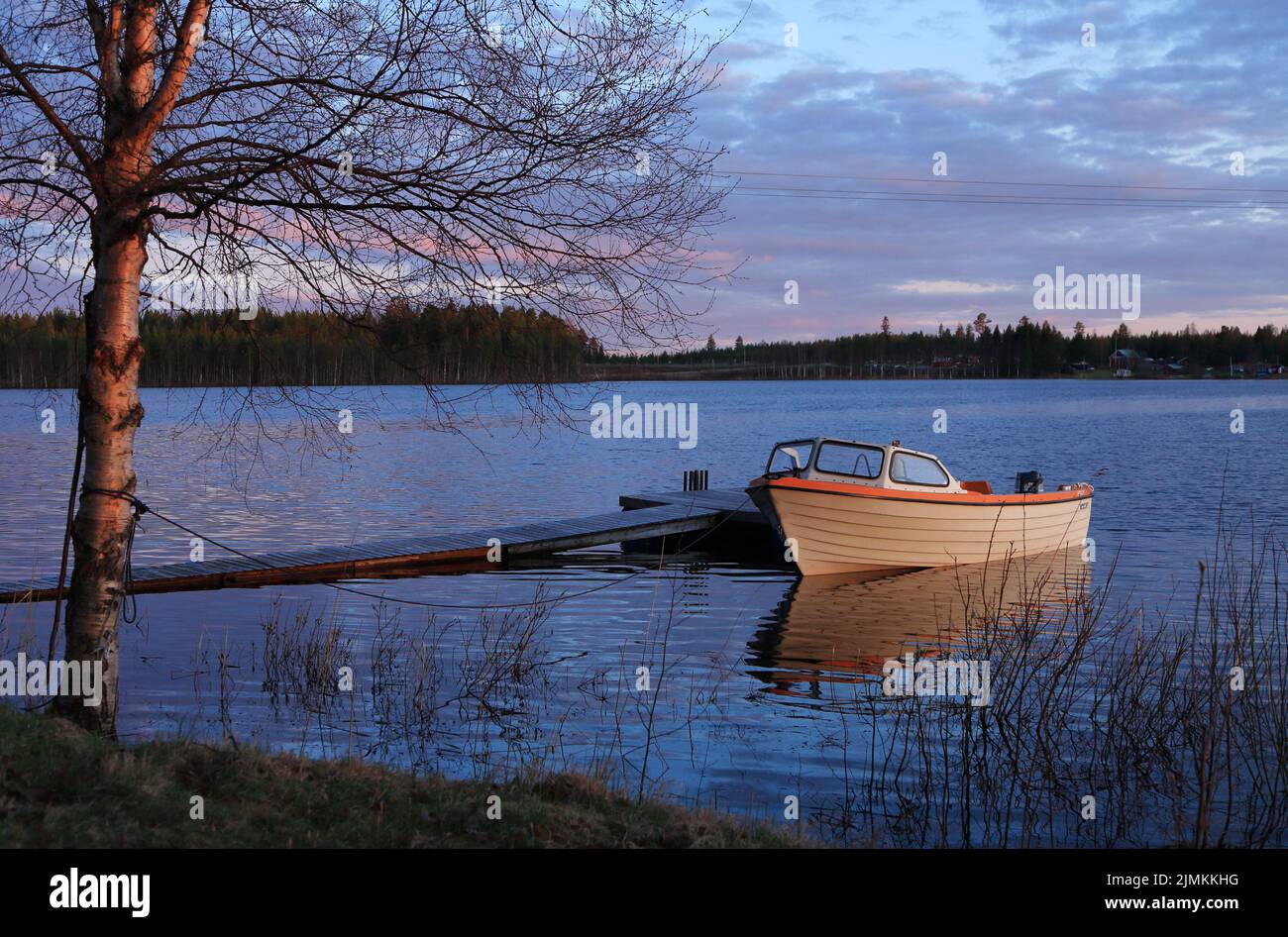 Noche de primavera con puesta de sol junto al lago en el norte de Suecia, donde un pequeño barco a motor con parte superior abierta de plástico para uso recreativo está amarrado en el embarcadero. Foto de stock
