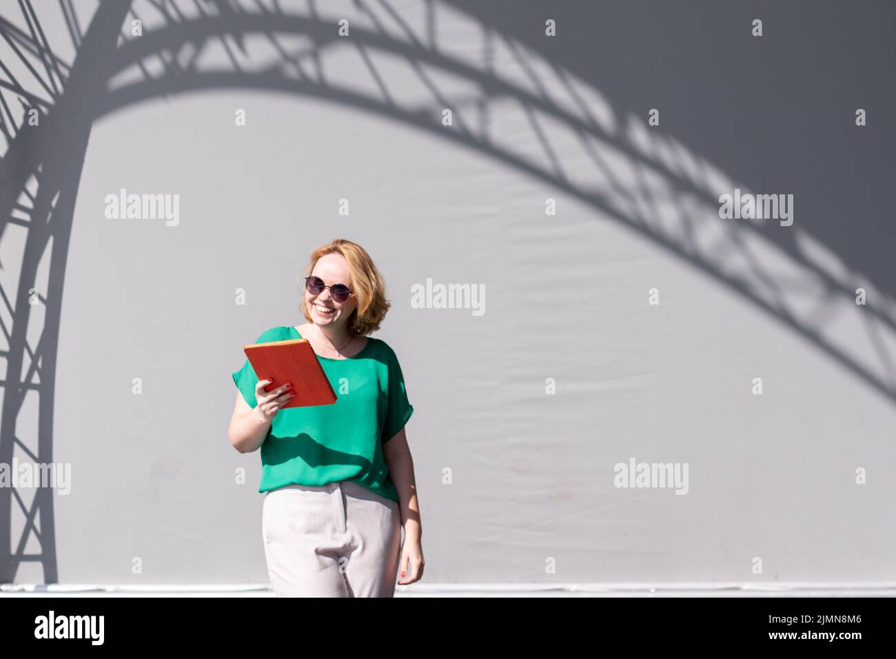 Chica feliz en gafas de sol hojeando la tableta, apoyado en la pared en la calle, hermosa sombra. Aprendizaje a distancia, educación en línea y trabajo Foto de stock