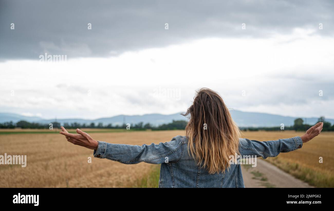 Vista desde atrás de una joven mujer caucásica en chaqueta vaquera de pie en la hermosa naturaleza de los campos de trigo dorado con sus brazos extendidos ampliamente. Foto de stock