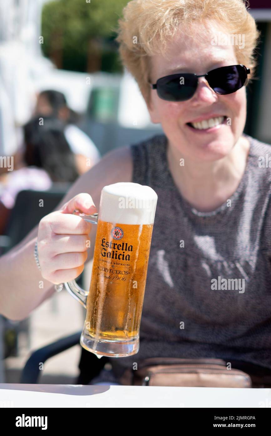 Una cliente sentada en una mesa fuera de un bar. Ella está sentada a la luz del sol y sosteniendo un vaso grande de lager Estrella Galicia. Foto de stock