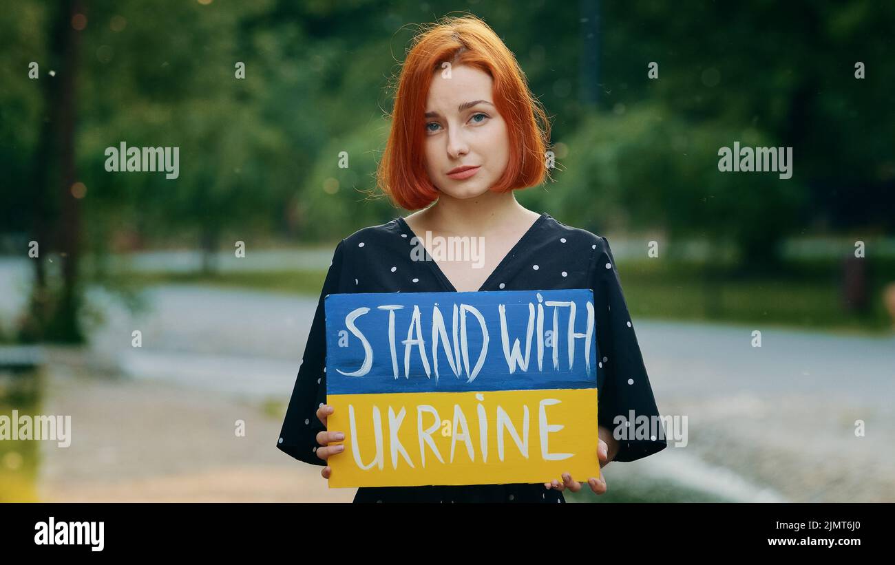 Triste pelirroja niña sostener cartón con inscripción escrita soporte de frase con Ucrania muestra bandera patriótica pintada en amarillo azul nacional ucraniano Foto de stock