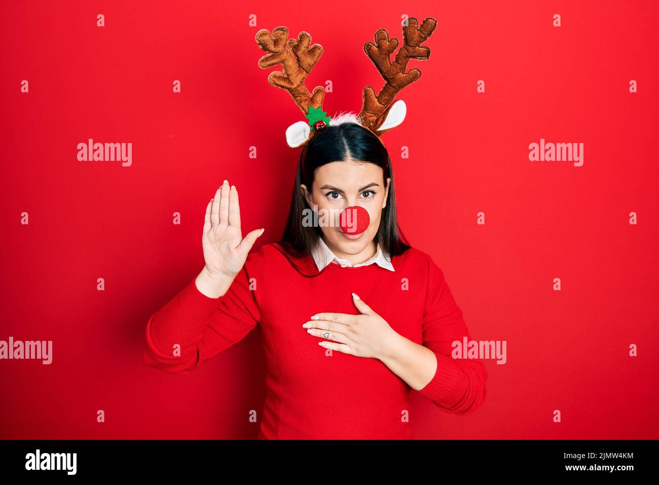 Mujer joven hispana con sombrero de Navidad de ciervo y nariz roja jurando con la mano en el pecho y la palma abierta, haciendo un juramento de promesa de lealtad Foto de stock