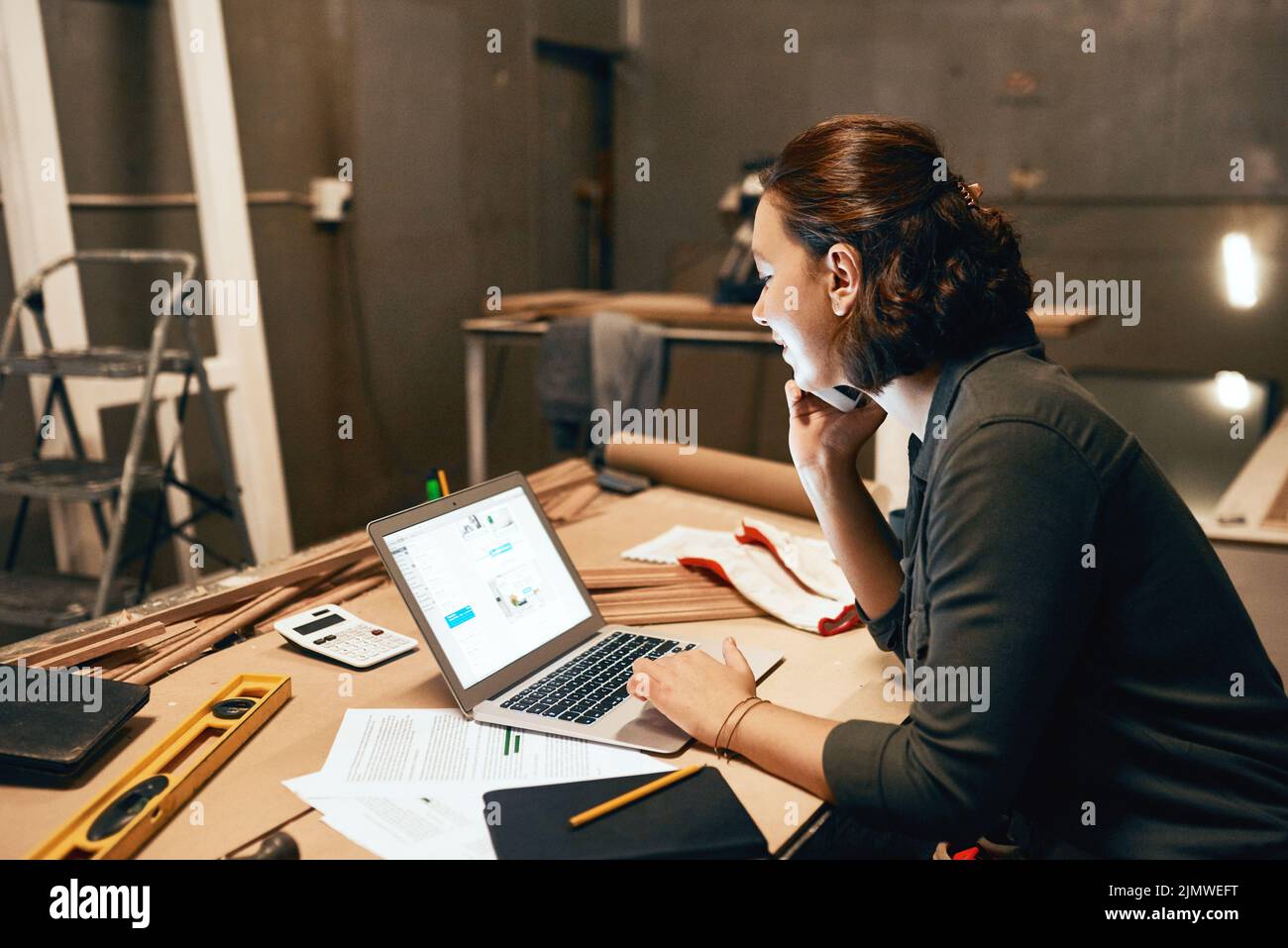 Tengo algunas cosas entre las que elegir. Una joven y atractiva carpintera trabajando en su portátil mientras habla por su teléfono móvil dentro de un taller. Foto de stock