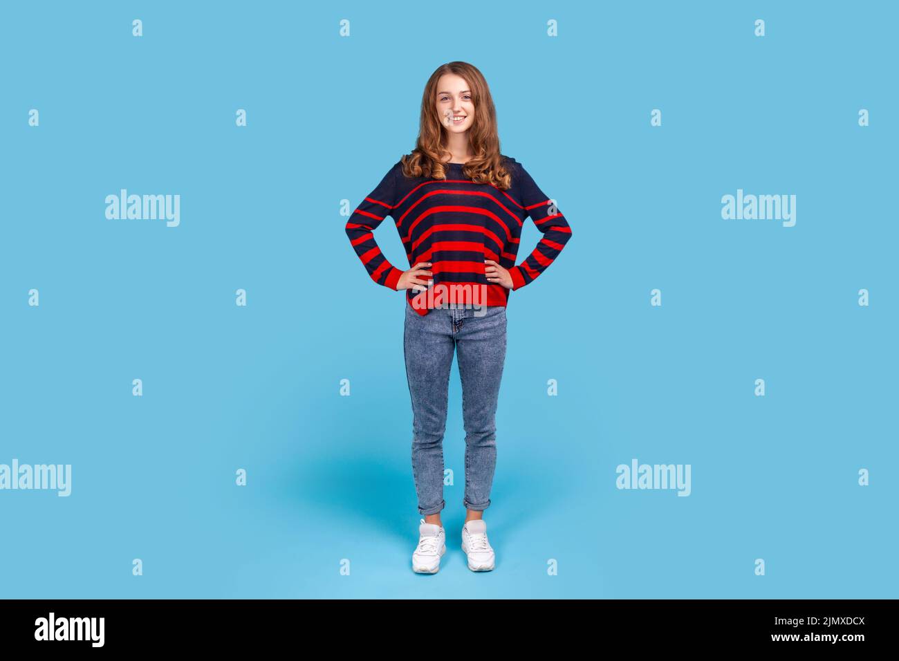 Retrato completo de una mujer feliz positiva con jersey de rayas y jeans de pie con las manos en las caderas mirando a la cámara. Estudio de interior aislado sobre fondo azul. Foto de stock