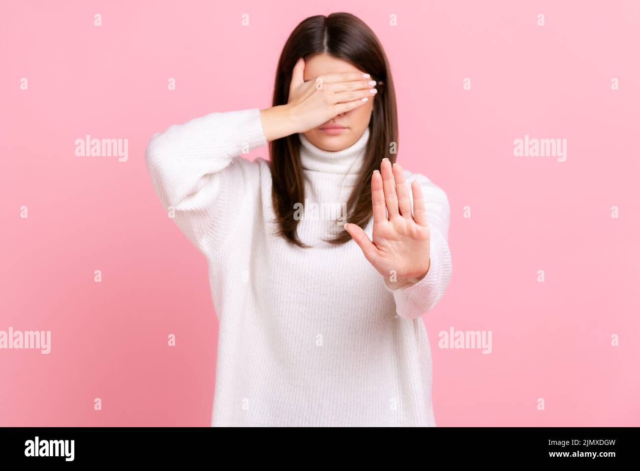 Mujer confusa cubre los ojos, levantando la mano para parar, sintiéndose estresado, negándose a ver contenido de miedo, usando suéter blanco de estilo casual. Estudio de interior grabado aislado sobre fondo rosa. Foto de stock