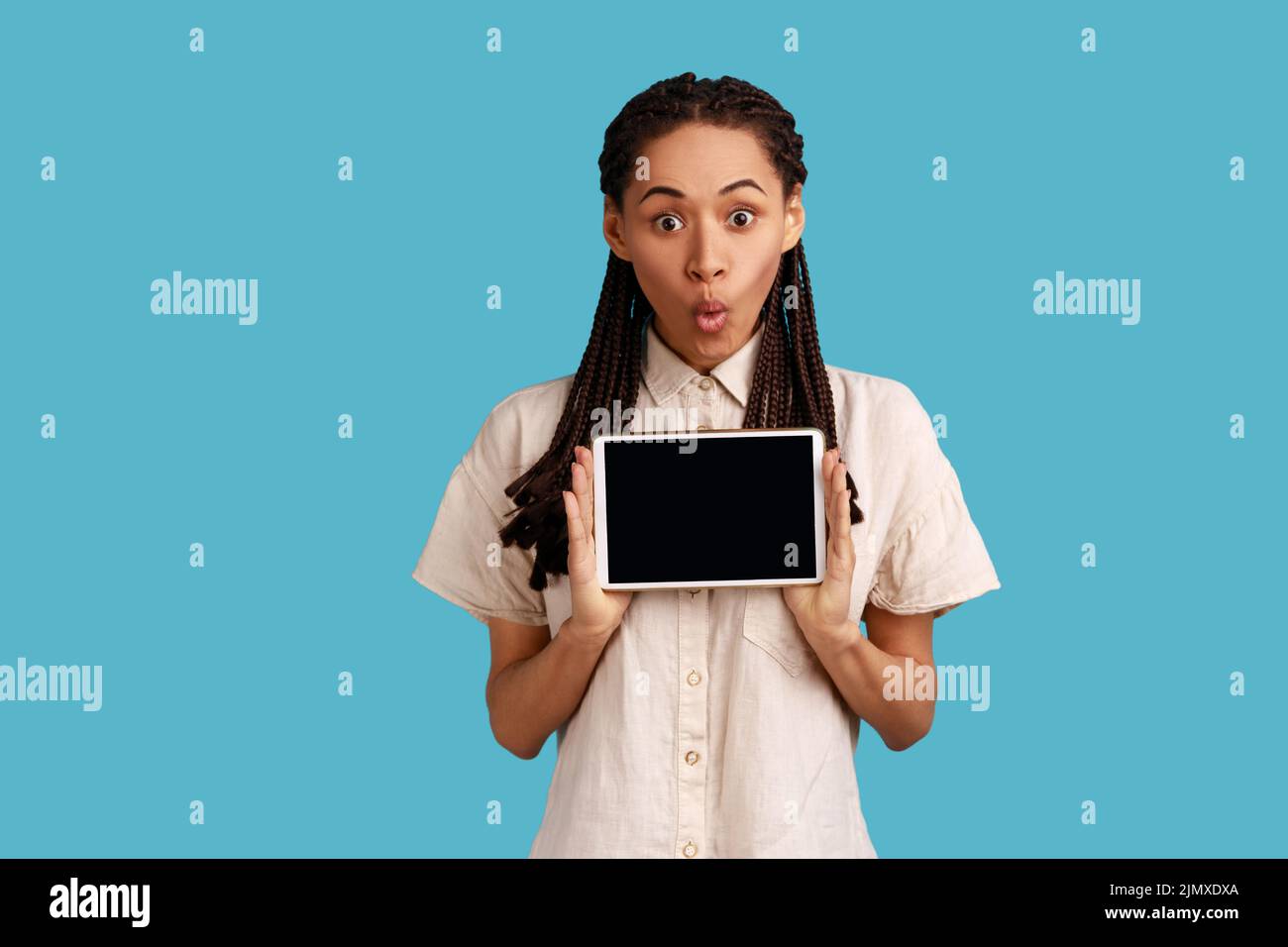 Asombrado sorprendido mujer sostiene tableta moderna con pantalla vacía para su texto promocional, posa con dispositivo electrónico en las manos, usando camisa blanca. Estudio de interior aislado sobre fondo azul. Foto de stock