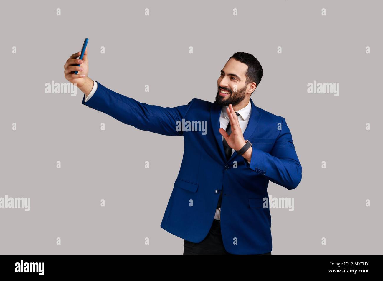 Hombre haciendo selfie en la cámara del smartphone, blogger comunicando, grabación de vídeo para los seguidores, agitando la mano, saludando, llevando traje de estilo oficial. Estudio de interior grabado aislado sobre fondo gris. Foto de stock