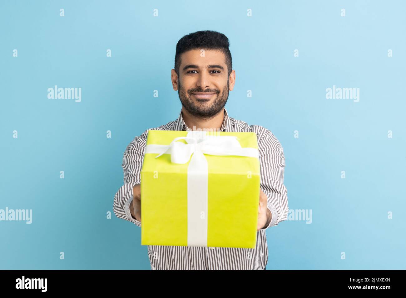 Retrato de un hombre de negocios barbudo sonriente con caja de regalo amarilla envuelta y sonriendo con cámara fotográfica, feliz día de fiesta, usando camisa a rayas. Estudio de interior aislado sobre fondo azul. Foto de stock