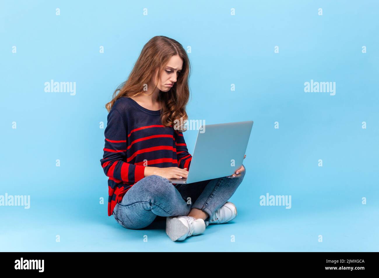 Retrato de una mujer molesta con un jersey a rayas de estilo informal, sentada sosteniendo el cuaderno de notas sobre las rodillas, mirando la pantalla, sintiendo tristeza. Estudio de interior aislado sobre fondo azul. Foto de stock